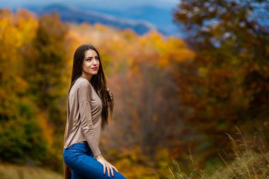 Uzun saçlı ve ince vücutlu genç bir esmer sonbahar manzarasında poz veriyor. .