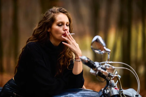一位美丽的长发女子骑着摩托车在林荫大道上抽烟 — 图库照片