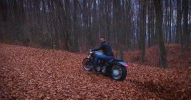 Sonbaharda yapraklarla kaplı bir yolda özel yapım kafe yarış motosikletine binen deri ceketli beyaz erkek motorcu. 4K UHD