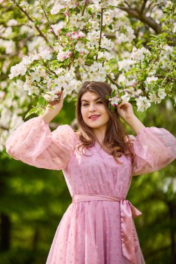 a young woman in a pink dress posing next to a blossoming apple, spring portrait.