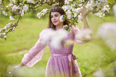 Pembe elbiseli genç bir kadın çiçek açan bir elmanın yanında poz veriyor, bahar portresi..