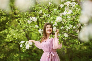 Pembe elbiseli genç bir kadın çiçek açan bir elmanın yanında poz veriyor, bahar portresi..
