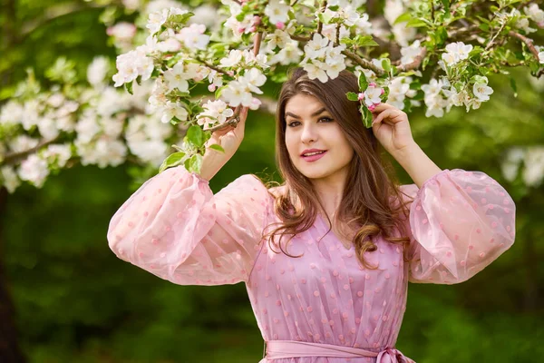 Pembe elbiseli genç bir kadın çiçek açan bir elmanın yanında poz veriyor, bahar portresi..