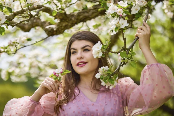 Pembe elbiseli genç bir kadın çiçek açan bir elmanın yanında poz veriyor, bahar portresi..