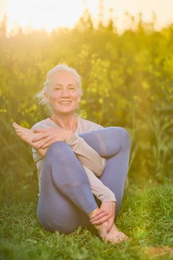 Yoga egzersizi, sabah rutini, gün batımı ışığı ve açık havada yeşil çimlerde dinlenen yaşlı bir kadın. Spor giyim sektöründe olgun bir kadın açık havada esneme hareketleri yapıyor. Yaş, sağlıklı yaşam tarzı.