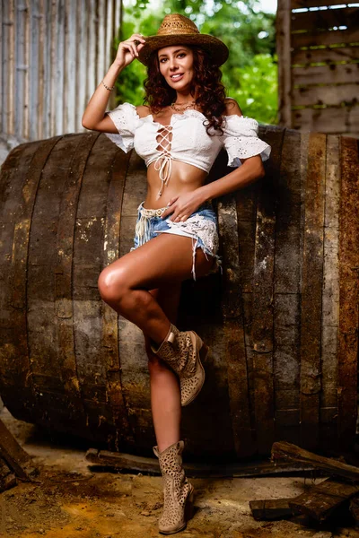 Woman in straw cowboy hat near old wooden barrel on brown wooden wall background.