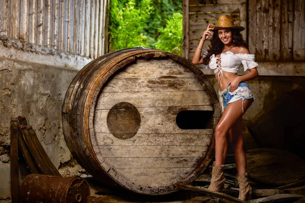 Woman in straw cowboy hat near old wooden barrel on brown wooden wall background.