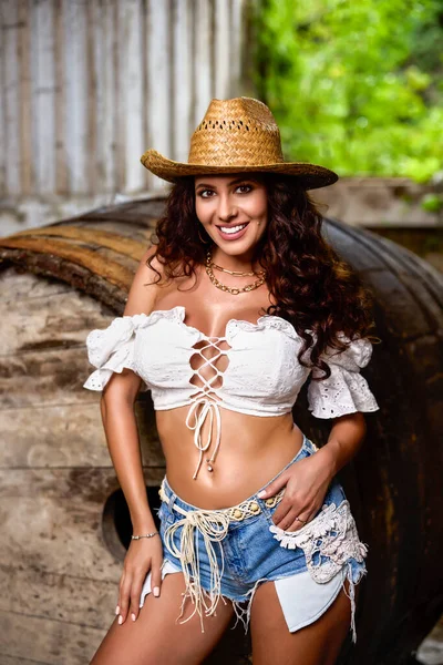 Woman in straw cowboy hat near old wooden barrel on brown wooden wall background.