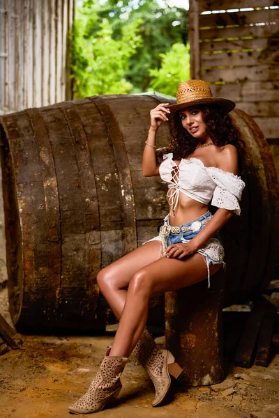 Woman in straw cowboy hat near old wooden barrel on brown wooden wall background.