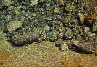 Çakıl taşlarıyla dolu büyük bir kaya havuzunda dalgalar ve güneş ışığı refleksleri