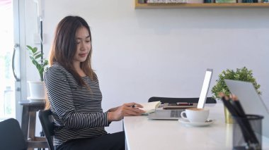 Genç Asyalı kadın kitap okuyor ve kafede dizüstü bilgisayar kullanıyor..