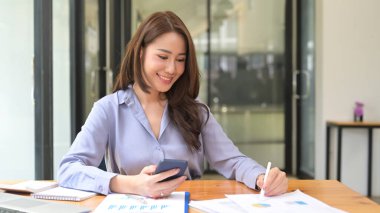 Gülümseyen iş kadını elinde akıllı bir telefon tutuyor ve mali belgeleri okuyor..