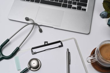 Doktor çalışma alanının üstünde dizüstü bilgisayar, dosya, stetoskop ve kahve fincanı var..
