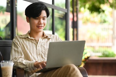 Gülümseyen Asyalı genç adam açık kafede oturuyor ve bilgisayar laptopu kullanıyor..