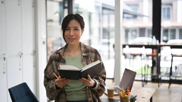 現代の職場で立ちながら本を持ちカメラに微笑む若い女性 — ストック写真
