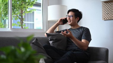 Gülümseyen Asyalı adam akıllı telefonda konuşuyor ve oturma odasında kanepede oturuyor..