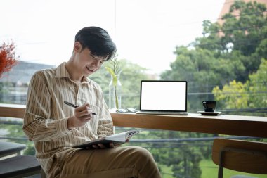 Gülümseyen Asyalı serbest çalışan modern kahve dükkanında oturuyor ve deftere yeni fikirler yazıyor..