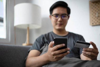 Gülümseyen Asyalı adam akıllı telefon kullanıyor ve internetten alışveriş yapmak için kredi kartı tutuyor..
