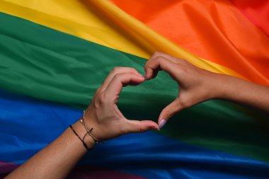 LGBT kadınlarının elleri bir arada ve gökkuşağı bayrağı üzerinde kalp şekli oluşturuyorlar. LGBT gururu kavramı.