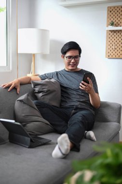 Genç Asyalı adam koltukta oturuyor ve akıllı telefon kullanıyor..