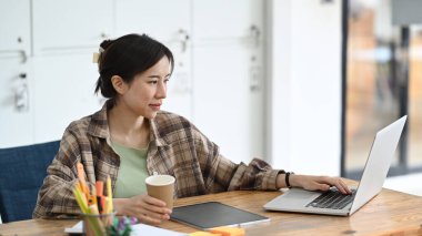 Güzel bayan tasarımcı elinde kahve fincanı tutuyor ve yaratıcı ofiste bilgisayar laptopuyla çalışıyor..