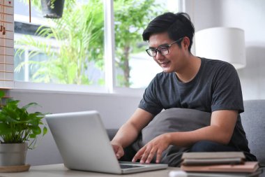 Genç Asyalı adam rahat koltukta oturuyor ve bilgisayar laptopu kullanıyor..