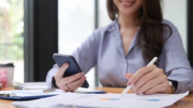 Gülümseyen kadın muhasebeci akıllı telefonu tutuyor ve finansal belgeleri analiz ediyor..