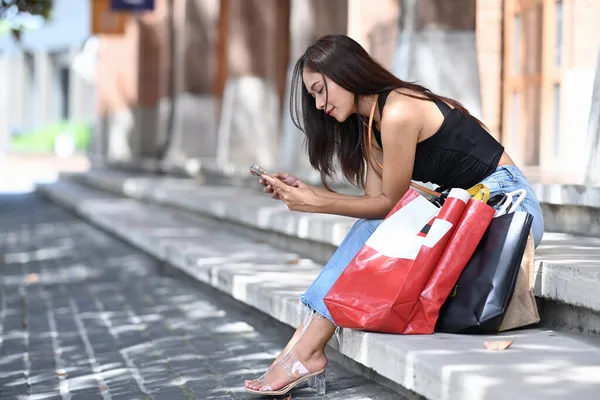 Dışarıda Alışveriş Torbalarıyla Oturan Akıllı Telefon Kullanan Genç Bir Kadın — Stok fotoğraf