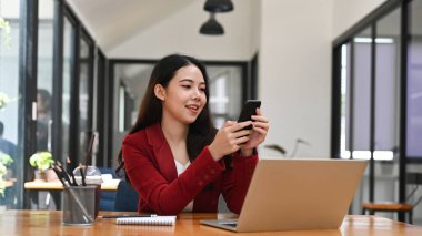 Asyalı iş kadını dizüstü bilgisayarın önünde oturuyor ve akıllı telefon kullanıyor..
