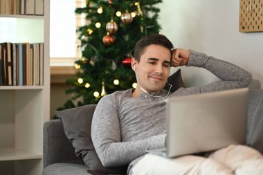 Kulaklık takan ve koltukta dizüstü bilgisayar kullanan gülümseyen adam..