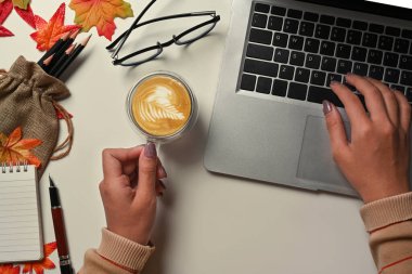 Elinde bir fincan kahve tutan ve sonbahar akçaağaç yapraklı beyaz masada dizüstü bilgisayar kullanan bir kadın..