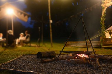 Akşamları kamp çadırının yanında şenlik ateşi ve arka planda oturan bir grup insan..