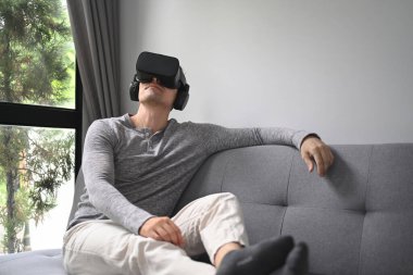 Genç adam evdeki kanepede sanal gerçeklik kulaklığıyla film izliyor..