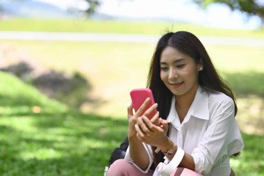 Parkta otururken akıllı telefon kullanan gülümseyen Asyalı genç kadın..