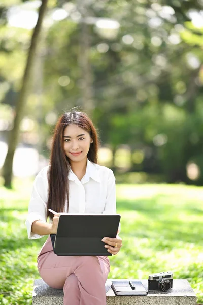 照片中的年轻女子坐在公园的长椅上 手持数码平板电脑 对着相机微笑 — 图库照片