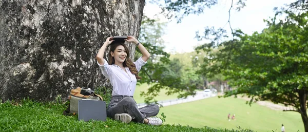 年轻的亚洲女人坐在树下的绿草上 微笑着对着相机 — 图库照片