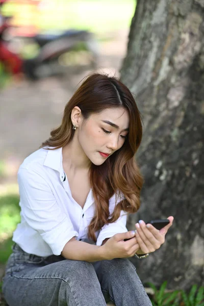 年轻的亚洲女人坐在公园的绿草上 一边用智能手机 — 图库照片
