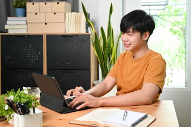 Günlük giysiler içinde gülümseyen Asyalı adam evde bilgisayar tabletiyle internette geziniyor..