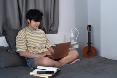 Gülümseyen Asyalı adam yatak odasında dizüstü bilgisayarla internette geziniyor..