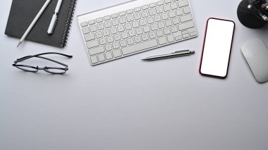 Modern workspace with smart phone, wireless keyboard and glasses on white table.