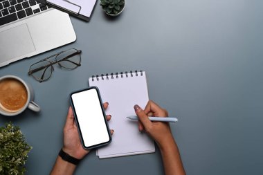 Overhead view woman holding smart phone and making notes on notebook.