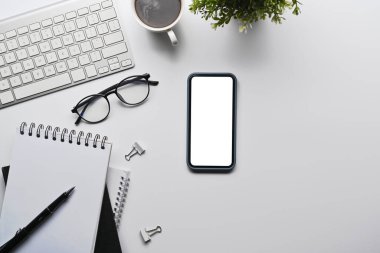 Top view smart phone, eyeglasses and notebook on white office desk.