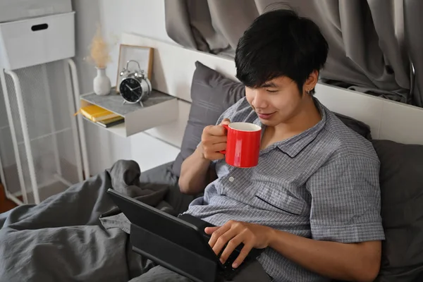 Szczęśliwy Azji Człowiek Picia Kawy Surfing Internet Komputer Tablet Łóżku — Zdjęcie stockowe