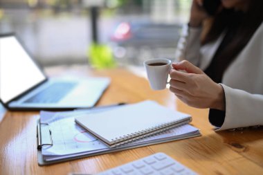 Kırpılmış iş kadını elinde kahve fincanıyla ofiste dizüstü bilgisayarla çalışıyor..