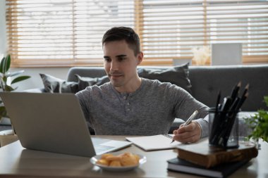 Gülümseyen adam dizüstü bilgisayar kullanıyor ve not defteri üzerinde not alıyor..