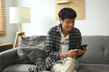 Gülümseyen Asyalı adam kanepede oturuyor ve internetten alışveriş yapmak ya da sosyal medyayı kontrol etmek için akıllı telefon söylüyor..