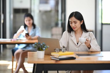 Genç bayan ofis çalışanı kahve içiyor ve bilgisayardaki e-postaları kontrol ediyor..