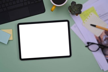 Üst görünüm dijital tablet, kahve fincanı, yapışkan notlar ve yeşil zemin üzerine dökümanlar.