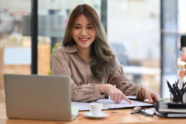 Dizüstü bilgisayarda çalışan ve ofis masasında doküman okuyan çekici bir iş kadını..