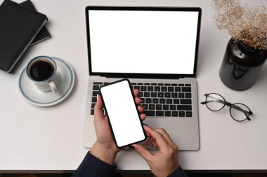 Yukarıda genç bir bayan dizüstü bilgisayarın önünde oturuyor ve ofis masasında akıllı telefon kullanıyor..
